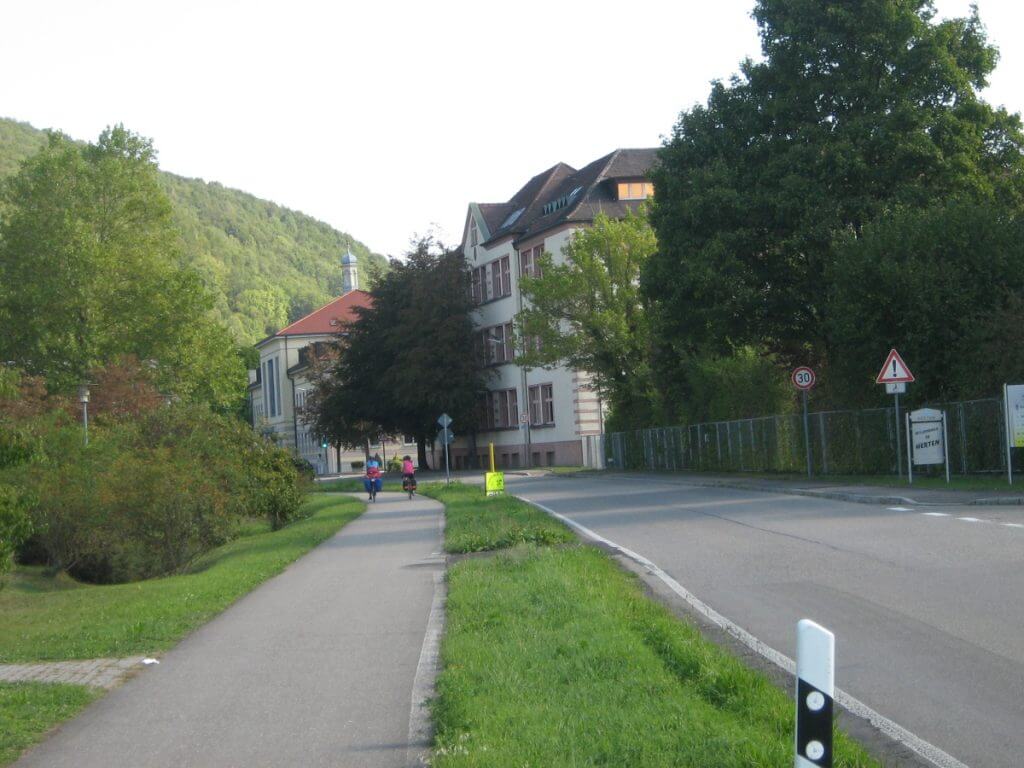 Cyclists enjoy a leisurely ride on the EuroVelo 15, Basel-to-Waldshut section, with a pathway that is nestled between lush greenery and a row of houses. The calm street, accompanied by traffic signs and the natural beauty of the hillside, offers a glimpse into the tranquil urban outskirts.