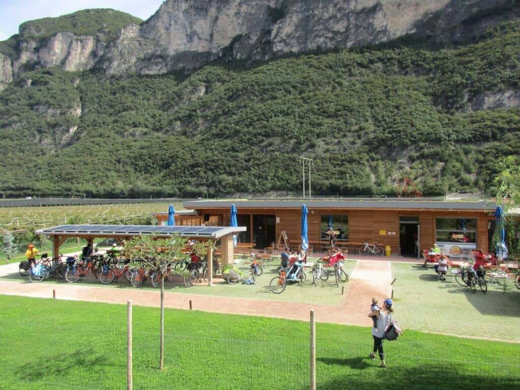 This image showcases a lively cyclist cafe along the Adige Bike route, nestled in a picturesque valley with towering cliffs and lush vegetation. The wooden structure of the cafe, complete with a covered seating area, is surrounded by parked bicycles, indicating its popularity among riders. Visitors are seen relaxing and enjoying refreshments in this outdoor setting