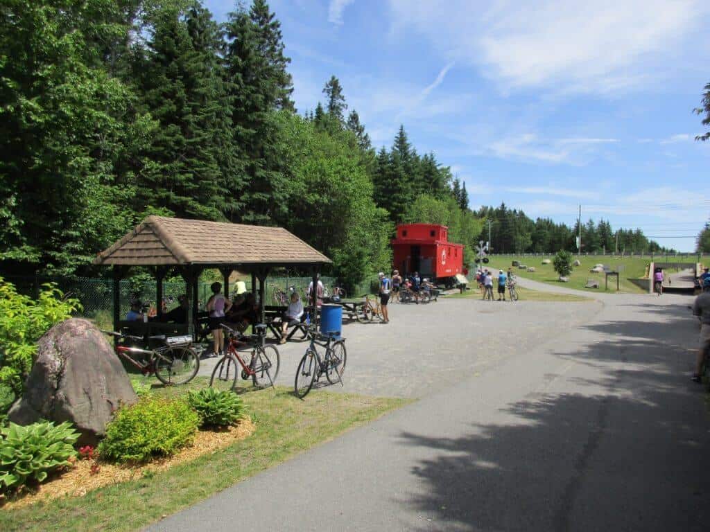 Cyclists and visitors rest at the shaded picnic area of the Corridor des Cheminots in Shannon, Quebec. A striking red caboose adds historical charm to the rest stop, set against a backdrop of tall evergreens under a clear sky