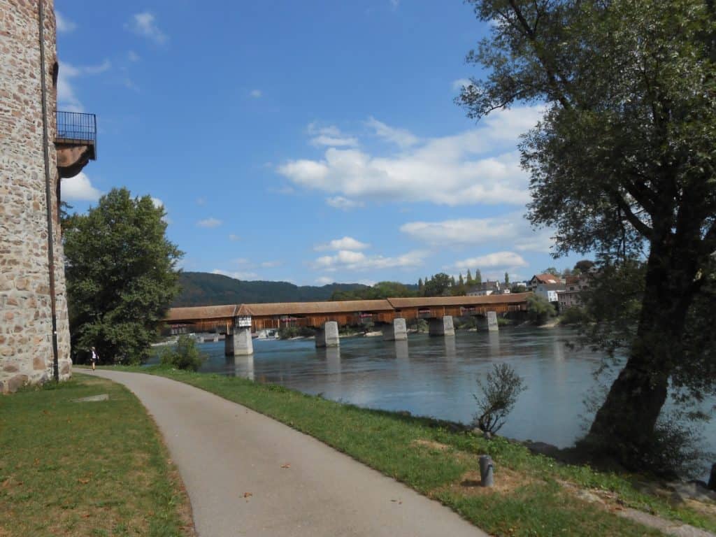 The Bad Säckingen Covered Bridge, a wooden structure spanning the Rhine River, is captured from a riverside path shaded by trees. The historic bridge connecting Germany and Switzerland is a testament to traditional architecture, set against a backdrop of a clear blue sky and the tranquil Rhine