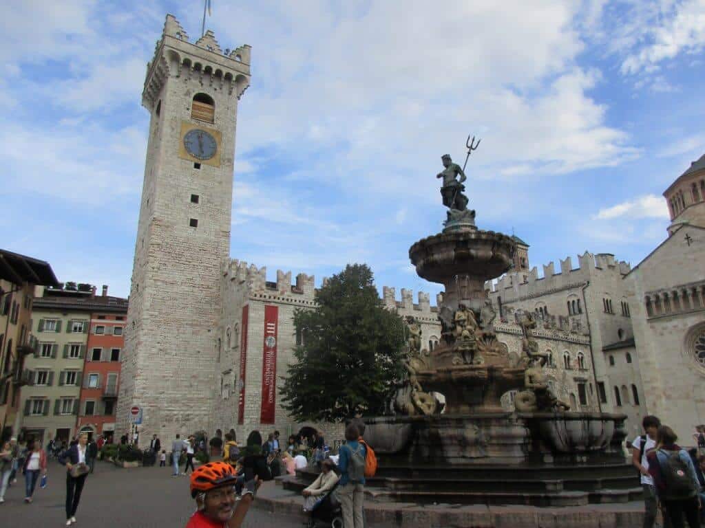 This image captures the vibrant atmosphere of the center of Trento, Italy, showcasing its historical architecture. A large, ornate fountain with a statue holding a trident dominates the foreground, while the imposing Torre Civica, a medieval tower with a clock, stands prominently in the background alongside other historical buildings.
