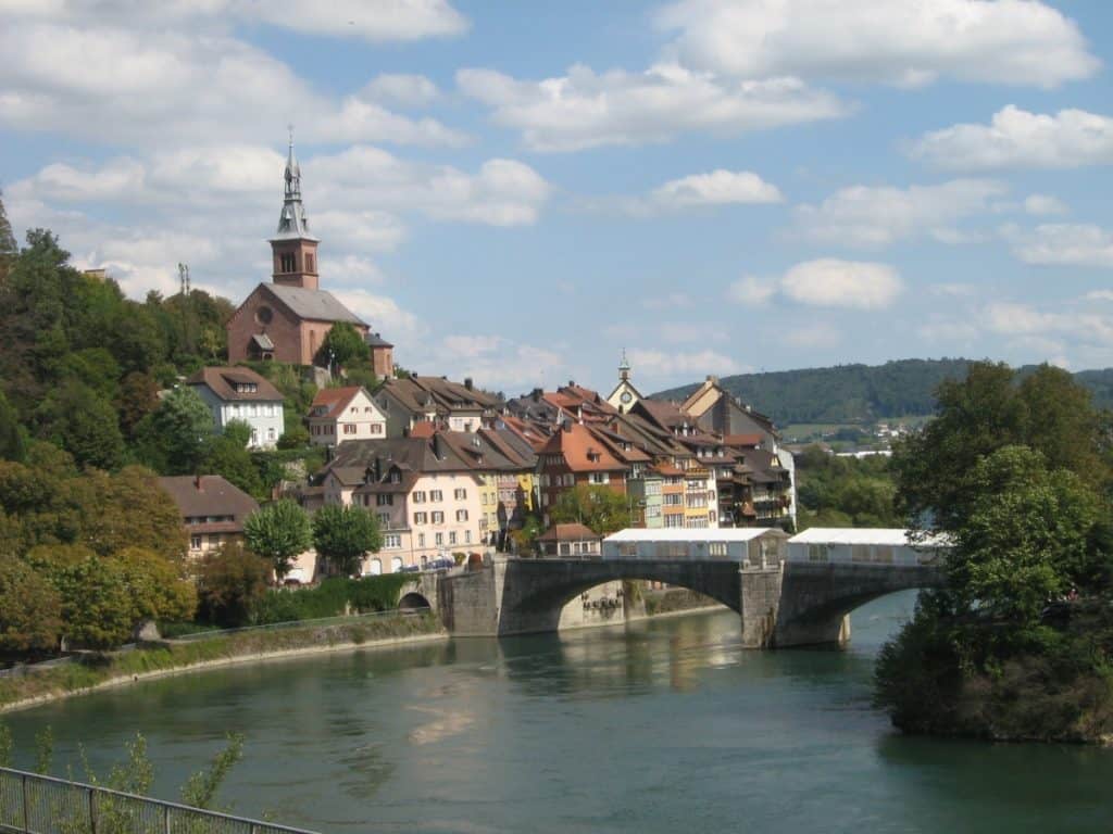 Laufenburg, a picturesque town divided by the Rhine River, viewed from the Rhine River Route. The town is characterized by its traditional architecture, including a prominent church with a red steeple, stone bridge, and houses with gabled roofs set against a backdrop of lush green hills