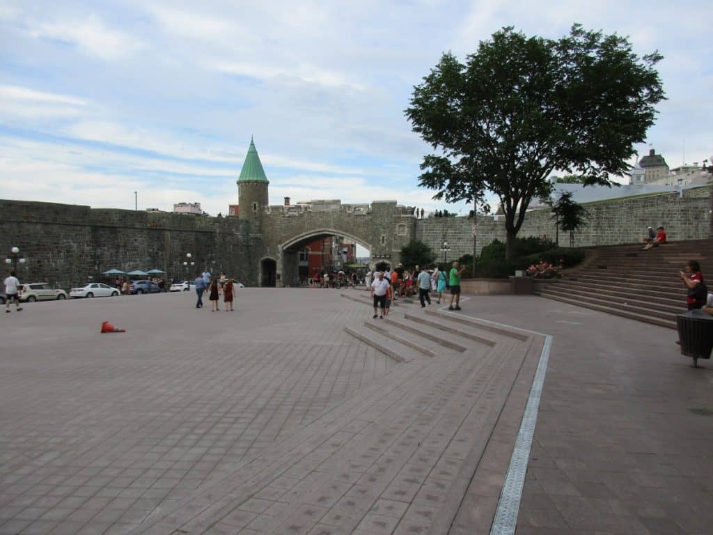 Visitors wander across the spacious Place d'Armes in Old Quebec, with the historic city gate and ramparts providing a sense of the area's rich past. The open square, lined with trees and benches, is a hub of activity against the backdrop of the iconic stone fortifications.