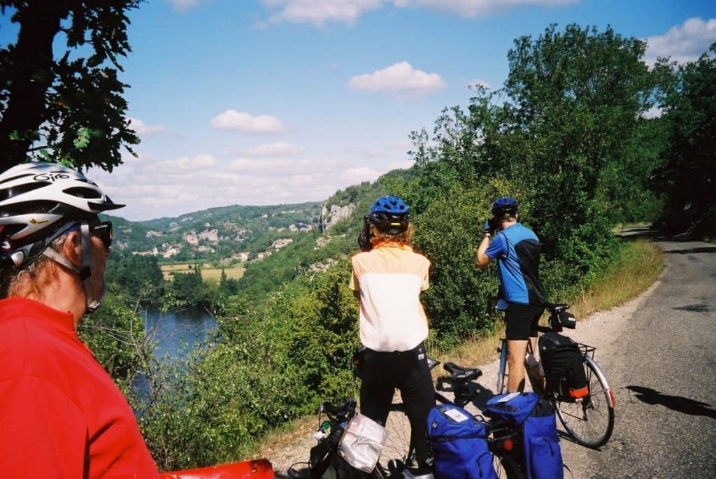 Cyclists with helmets and equipped bicycles stand on a roadside, overlooking the scenic River Lot valley in France, with lush greenery and cliffs under a partly cloudy sky.