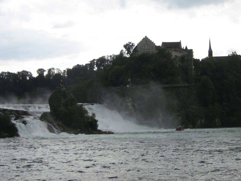 The majestic Rhein Falls in Switzerland, where cascading waters create a misty atmosphere against the backdrop of a medieval castle perched atop a lush cliff. A small boat braves the turbulent waters near the falls, offering a sense of scale to this natural wonder under an overcast sky