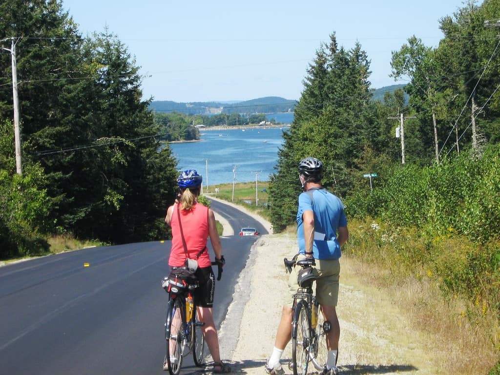 Two cyclists, one in red and one in blue, standing beside their bikes, enjoy the coastal views on a sunny day while cycling on Deer Isle, Maine, with a scenic overlook of the blue waters and distant land.