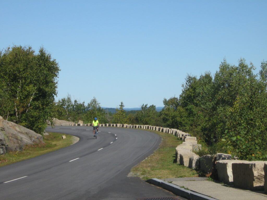A cyclist in a bright yellow shirt rides along a winding road in Acadia National Park, Maine, surrounded by lush greenery and a clear blue sky.