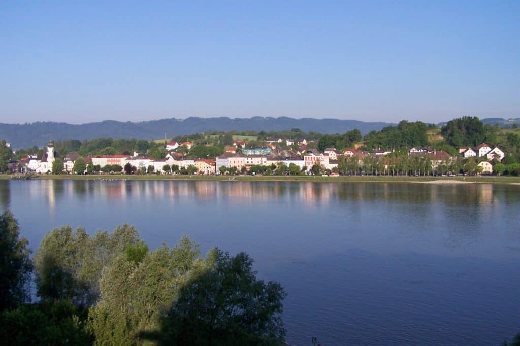 Panoramic view of Aschach, Austria, with its quaint buildings and church spires reflecting on the calm Danube River, flanked by lush greenery, under a clear, pale blue sky.