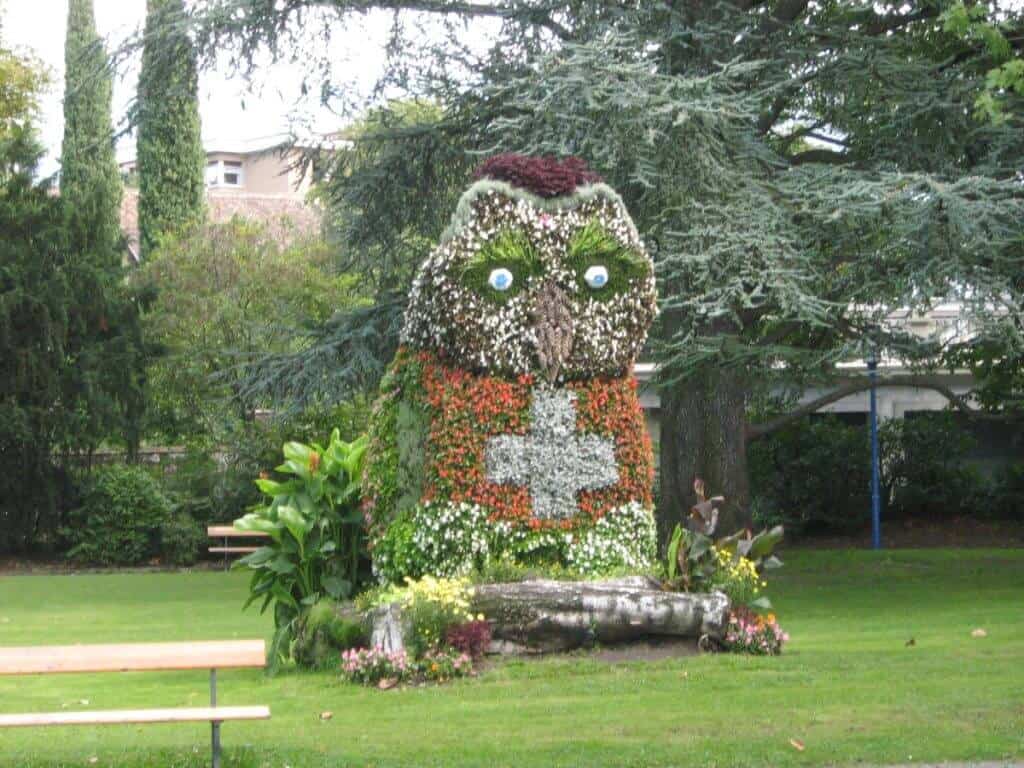 A creative floral display shaped like an owl, located in a park in Vevey, Switzerland. The owl is intricately designed with a variety of colorful flowers, making up its detailed feathers, expressive eyes, and distinctive beak. Surrounded by lush greenery and a wooden bench, the setting exudes a tranquil and artistic atmosphere