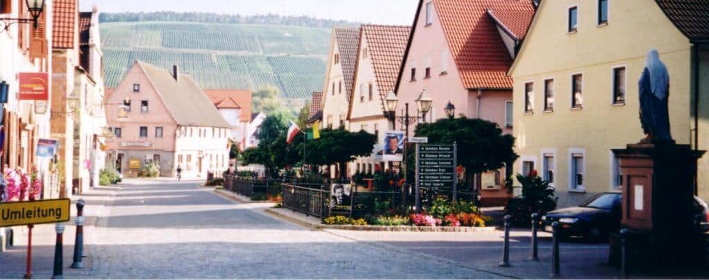 A peaceful street view in Markelsheim, Germany, showcasing a row of colorful houses along a tidy, paved road. The background reveals lush vineyards on the hills, enhancing the rural charm. Street signs, parked cars, and blooming flowers along the sidewalk contribute to the idyllic village atmosphere captured in this image.