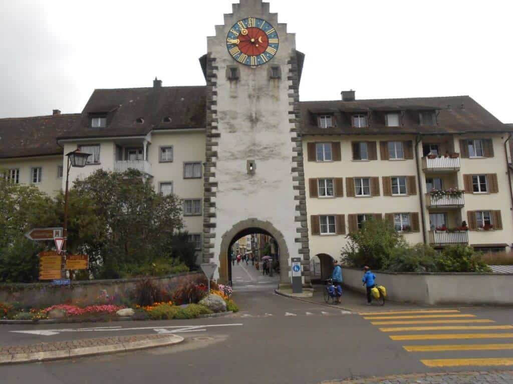 Cyclists prepare to pass beneath an archway in the historical tower of Stein am Rhein, Switzerland. The tower features a large clock face and is part of a building adorned with shutters and flower boxes, showcasing the quaint charm of this Swiss town.