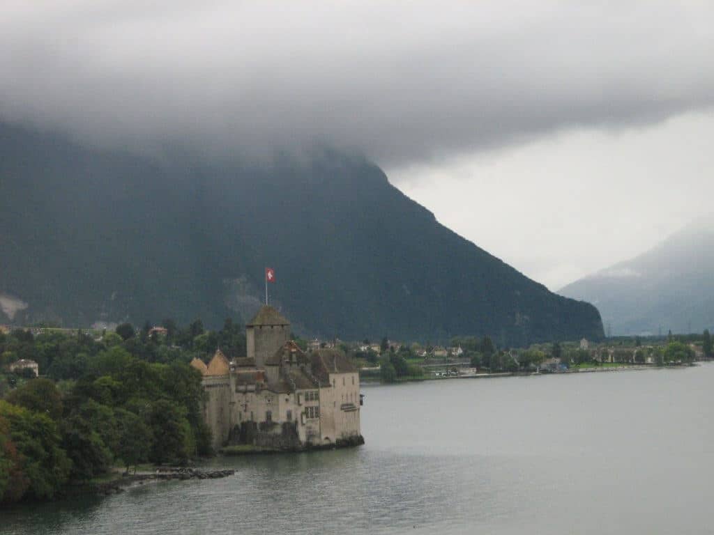 The image captures the iconic Château de Chillon, located on the shores of Lake Geneva near Montreux, Switzerland. The medieval castle, with its robust stone towers and walls, stands prominently on a small island connected to the mainland. Overcast skies and mist shroud the mountainous backdrop, adding a dramatic and mysterious atmosphere to this historic lakeside setting.