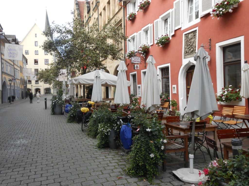The quaint outdoor seating of Hotel Pension Gretel in Konstanz, Germany, with wooden tables and folded parasols awaiting guests. Parked bicycles with touring gear hint at a popular resting spot for cyclists, amidst a cobblestone street lined with colorful, flower-adorned buildings