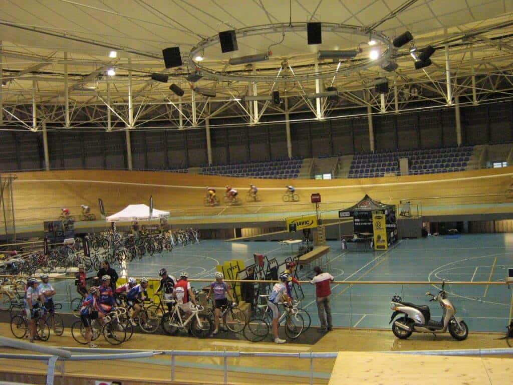 The interior of the World Cycling Center in Switzerland, featuring an active velodrome scene. Cyclists in various colorful outfits are seen racing on the track, while others prepare at the edge, next to rows of parked bicycles. The facility is equipped with overhead lights and surrounded by blue spectator seats, emphasizing its capacity for training and hosting competitive events. The atmosphere is dynamic, capturing the intensity and focus of competitive cycling.