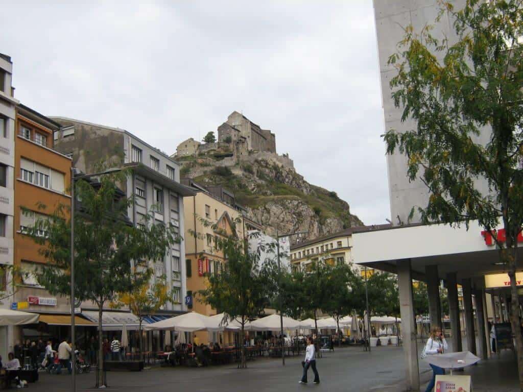 Bustling main street scene in Sion, Switzerland, with modern and traditional architecture blending seamlessly. In the foreground, a lively pedestrian area features cafes with outdoor seating, while colorful buildings line the street. Dominating the background is a striking, rocky hill topped by the ancient ruins of a castle, presenting a stark contrast between the urban environment and historical fortifications.