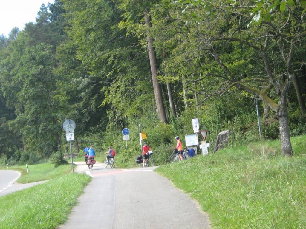 A group of cyclists tackle a gentle hill on the Bodensee Cycle Route, surrounded by lush greenery. Road signs for bicycles indicate a shared path, while a cyclist checks a map, suggesting a moment of navigation amidst the natural beauty of the route.