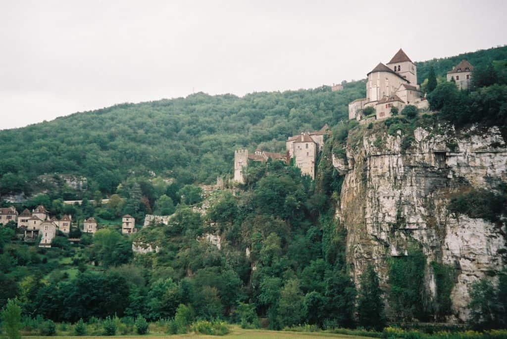 The medieval village of Saint Cirq Lapopie in France perched atop a steep cliff, with historic stone buildings nestled among green trees, showcasing the village's dramatic position above the landscape