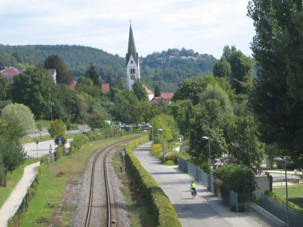 Cycling around Lake Constance along the Bodensee or Lake Constance bike route, capturing a cyclist in a bright green jacket pedaling on a paved path parallel to a railway track. The backdrop features a charming village with a prominent church spire rising above the treeline and green hills in the distance.