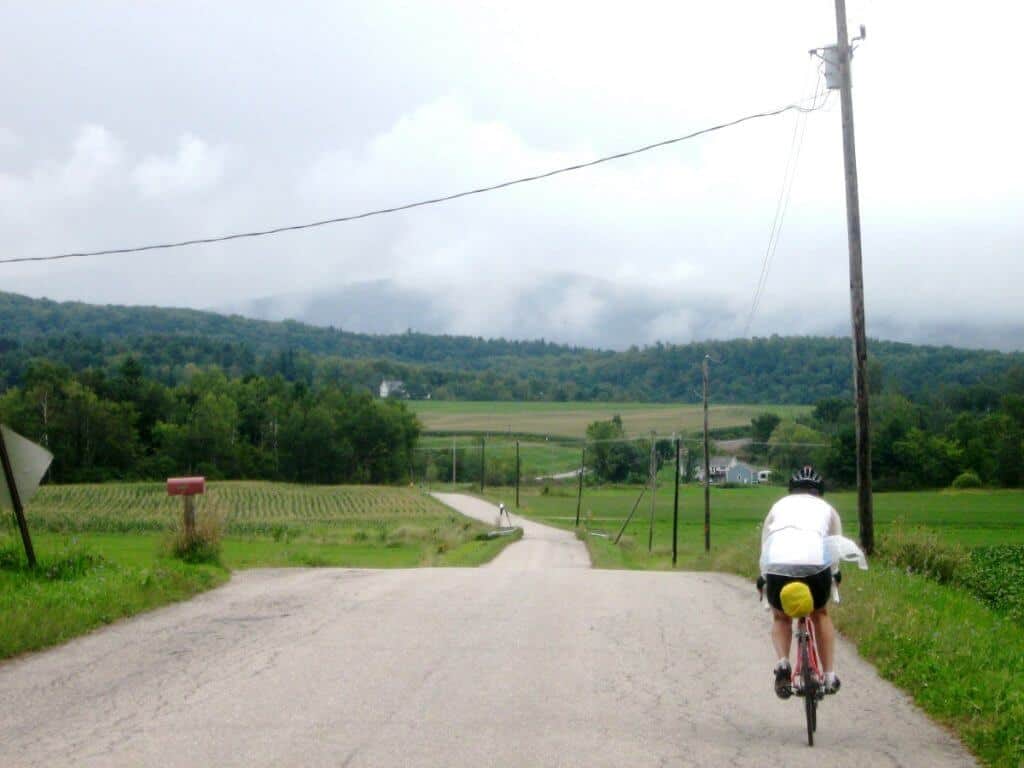 A cyclist in a white jersey and yellow shorts rides along Plank Road in Vermont, surrounded by expansive fields and rolling hills. The scene captures a serene rural landscape under a cloudy sky, with distant fog partially obscuring the green mountains.