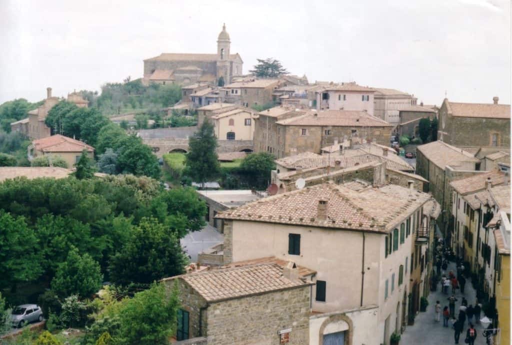 A view from above of the mountaintop village of Montalcino, Italy, showcasing the historic stone buildings with terracotta roofs and winding streets, with the prominent silhouette of a church against a hazy sky.