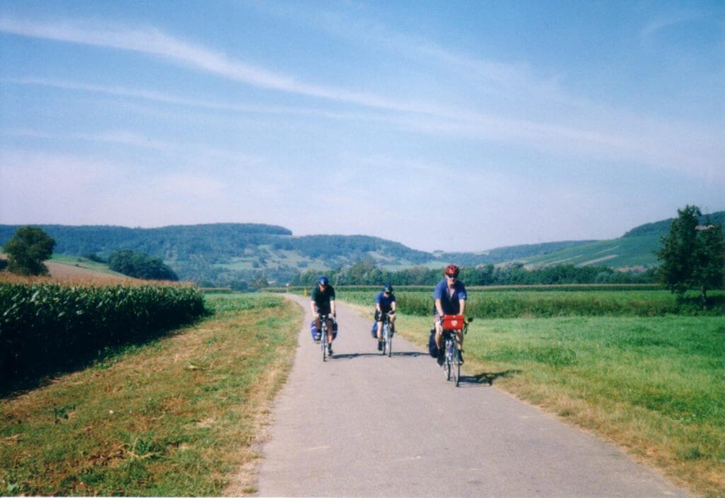 A serene image of three cyclists riding along the Romantic Road Bicycle Touring Route path surrounded by lush green landscapes. The path, bordered by fields and gentle hills, stretches under a clear blue sky with a few clouds. This captures a moment of leisure and exploration on what is known as the Romantic Road Bicycle touring route.