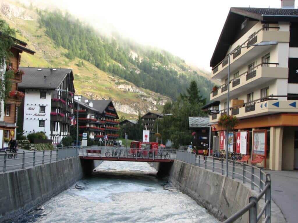 The image depicts a picturesque street in Zermatt, Switzerland, showcasing the town's charming architectural blend against a backdrop of misty mountains. A swift, clear river flows through a concrete channel in the center, bordered by pedestrian walkways and railings. Alpine-style buildings with balconies adorned with flowers line the street