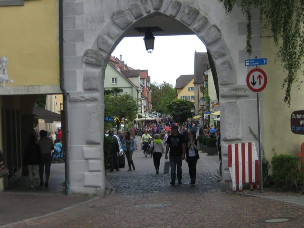 View through an old stone archway leading into the bustling main street of Meersburg, Germany. The arch frames a lively scene of pedestrians wandering along a cobblestone street lined with colorful buildings. The image captures the vibrant everyday life of the town, with people engaged in casual strolls