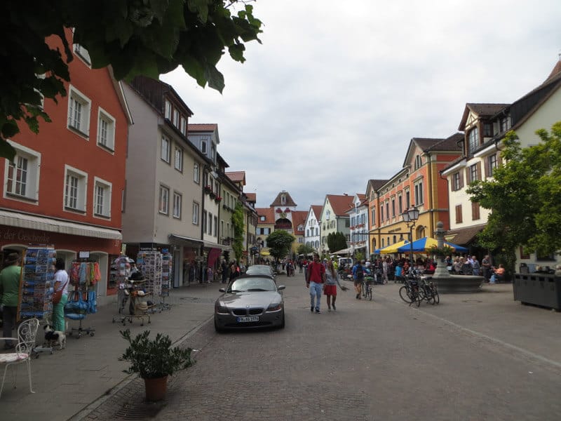 The bustling main street in Meersburg, Germany, where pedestrians and cyclists mingle among colorful facades of shops and cafes. The vibrant street life unfolds under a cloudy sky, with the charm of this historic town evident in its traditional architecture and inviting public spaces.