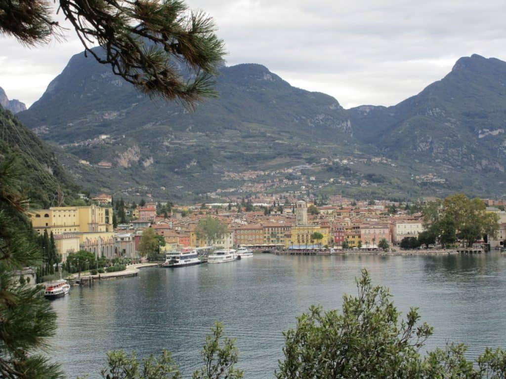 A picturesque view of the harbor of Riva del Garda, Italy, framed by pine branches in the foreground. The harbor is bustling with activity, featuring boats moored along the waterfront and a variety of colorful buildings lining the shore. The backdrop is dominated by towering mountains that rise steeply from the town, creating a dramatic contrast with the serene lake.