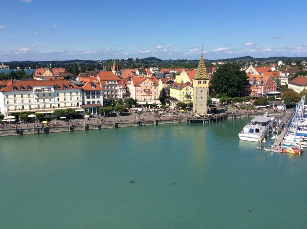 Aerial view of Lindau Harbor, Germany, showcasing the clear turquoise waters, moored boats, and the distinctive Mangturm tower amid colorful European buildings under a bright blue sky.