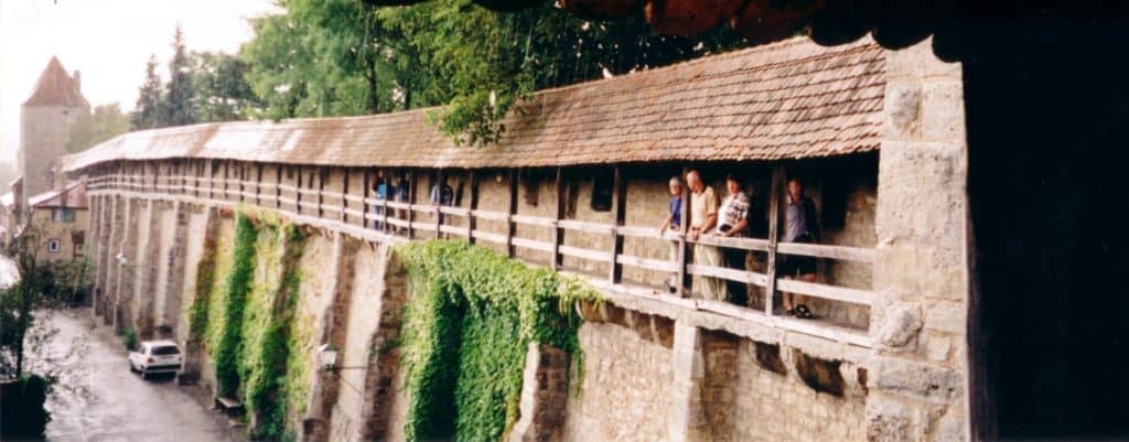 Visitors explore the historic covered walkway of the Rothenburg town walls in Germany, overlooking a street below. The wall is adorned with lush green ivy, enhancing its medieval architecture. This image captures a moment of cultural engagement and architectural beauty, with people peering from the walkway and a car passing beneath on the rain-slicked road.