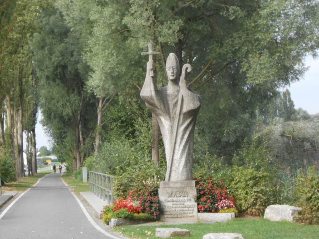 A statue of St. Pirmin, clutching a staff and a model of a church, greets visitors on a bicycle path leading to Reichenau Island in Germany. Cyclists can be seen in the distance traveling the tree-lined route, which is embellished with lush greenery and vibrant flower beds.