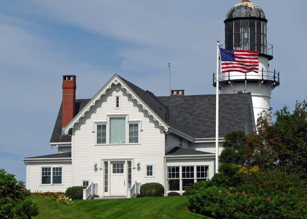 Cape Elizabeth Lighthouse in Maine, with its white, ornate structure and distinctive glass lantern room, adjacent to a classic New England-style home under a blue sky, complemented by an American flag waving proudly in the breeze.