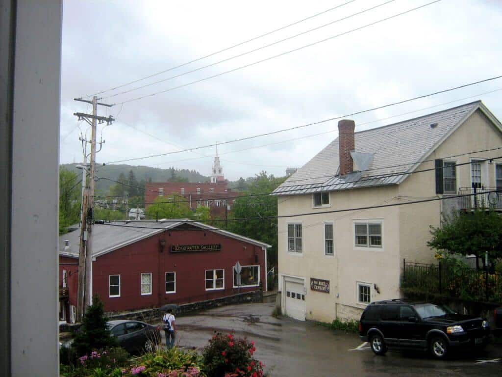 View of a rainy day in Middlebury, Vermont, showcasing local architecture and a lush green backdrop. The foreground features a parking lot with a few vehicles and a pedestrian with an umbrella, while the middle ground includes a red building labeled "Edgewater Gallery" beside a cream-colored building. In the background, the town's distinctive white church spire rises prominently against the overcast sky.