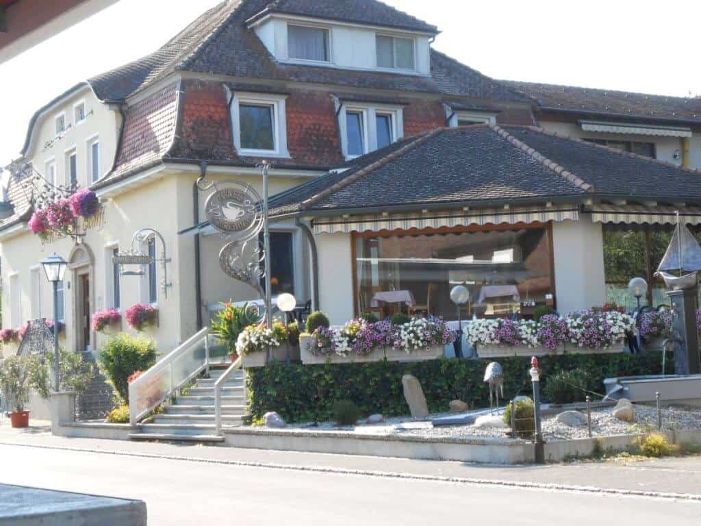 The front view of Gasthaus Schiff, a charming and inviting guesthouse in Moos, Germany, captured on a sunny day. The building is adorned with multiple flower boxes bursting with colorful blooms, enhancing the aesthetic of the traditional architecture with its quaint windows and red tiled roof. A sign with the establishment's name and a fish symbol hangs prominently, indicating its likely focus on seafood. The entry features a set of stairs leading up to a welcoming porch