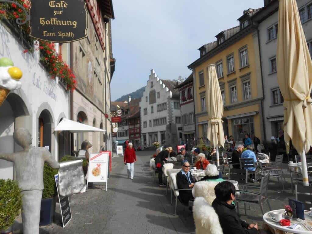 A lively street scene in Stein am Rhein, captured in front of Café zur Hoffnung. The café's outdoor seating is bustling with patrons enjoying their meals under umbrellas, while the street is lined with charming, historically rich buildings adorned with flower boxes. Statues and standing menus add a quaint touch to the cobblestone street, creating an inviting atmosphere for both locals and tourists. The background reveals the architectural beauty typical of this picturesque Swiss town.