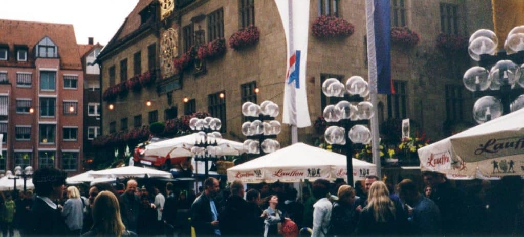 A bustling scene at the Heilbronn Wine Festival in Germany, showcasing a lively crowd of people gathered under white umbrellas branded with "Laurentia." The festival is set against a backdrop of historic buildings adorned with colorful flower baskets. Balloons and flags add a festive touch to the atmosphere, reflecting the lively spirit of this popular local event