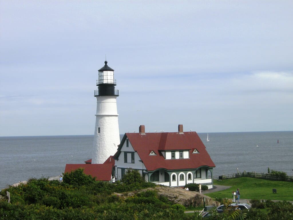 Iconic Portland Head Light in Maine, a historic white lighthouse with a black lantern room, next to a charming red-roofed keeper's house, perched on a grassy headland overlooking a serene ocean with sailing boats in the distance.