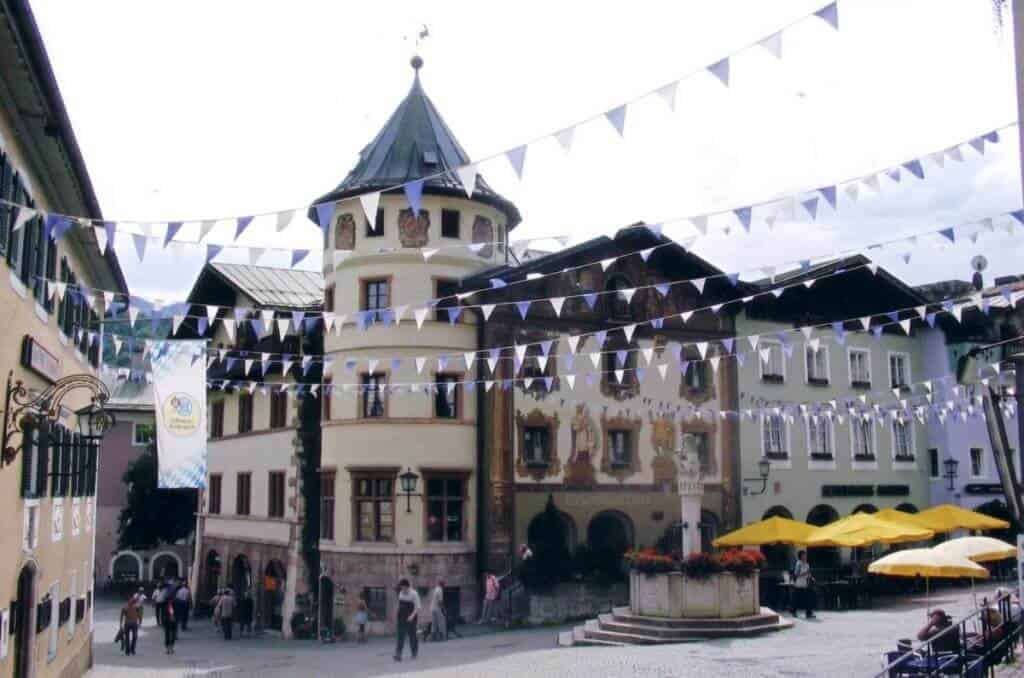 Festive atmosphere in Berchtesgaden, Germany, with triangular flags strung across the square, people milling about, traditional buildings, and a quaint tower, all set against a backdrop of a cloudy sky,