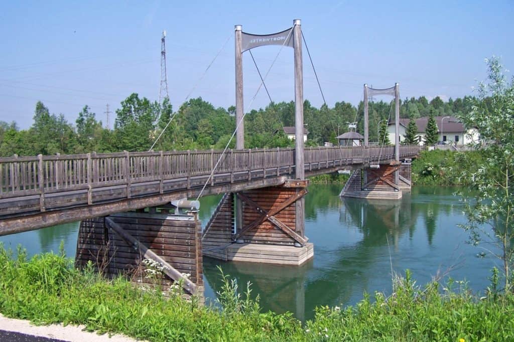 Europe's longest wooden bridge near Enns, Austria, spans a tranquil river with its sturdy timber structure and distinctive suspension cables, nestled in a serene natural setting with lush greenery.