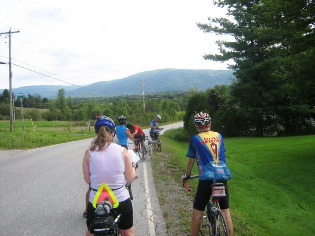 Bicycle touring in Vermont with a group of cyclists on a rural road near East Middlebury, Vermont. The cyclists, wearing various cycling gear, are riding along a paved road that cuts through a lush landscape with mountains visible in the background. A few cyclists are paused at the roadside, looking at the scenic, green mountainous view.