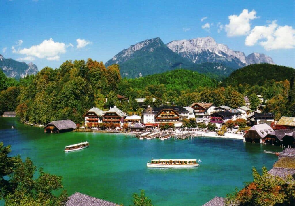 Vibrant scene at Königssee, Germany, featuring electric boats gliding on the emerald waters of the lake, surrounded by lush greenery and Alpine architecture, with the majestic Bavarian Alps towering in the background under a clear blue sky.