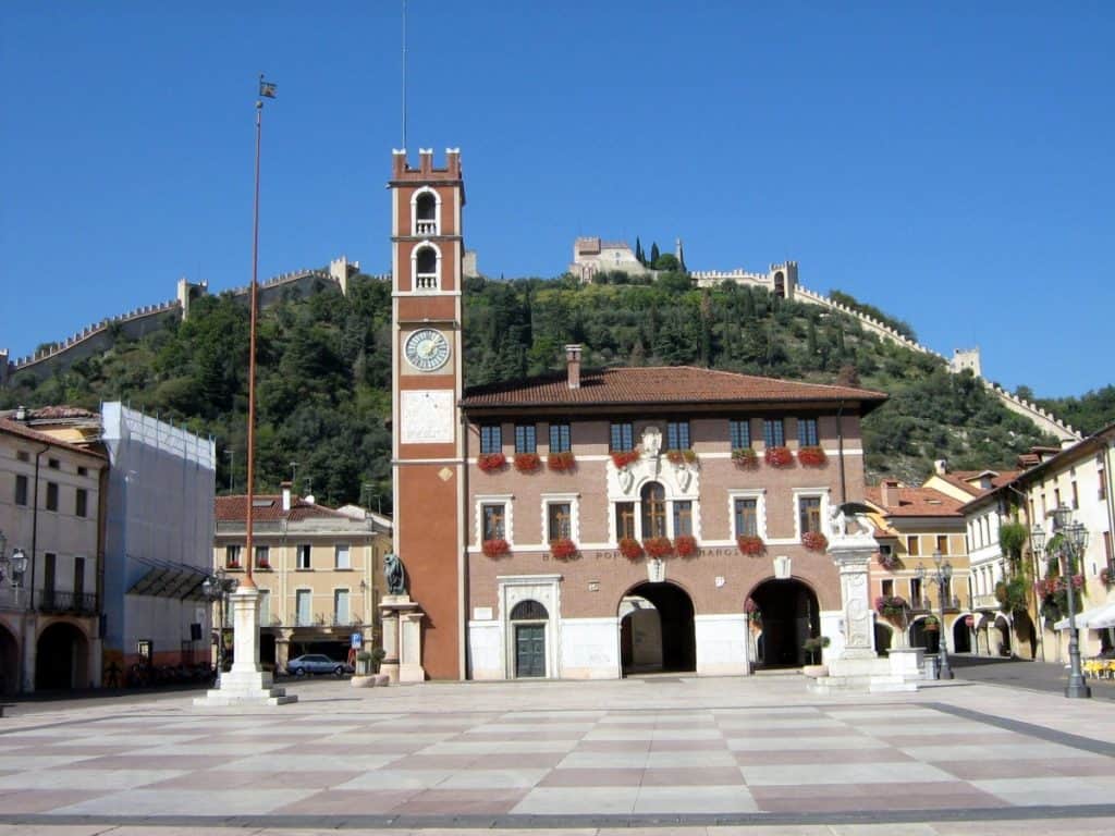 The town hall in the main square of Marostica, Italy, stands prominently with its red facade and clock tower against a clear blue sky. Behind it, the hillside is adorned with a well-preserved medieval wall that stretches across the landscape. The square is spacious and paved, evoking a sense of historical charm and readiness for the town's famous living chess match events.