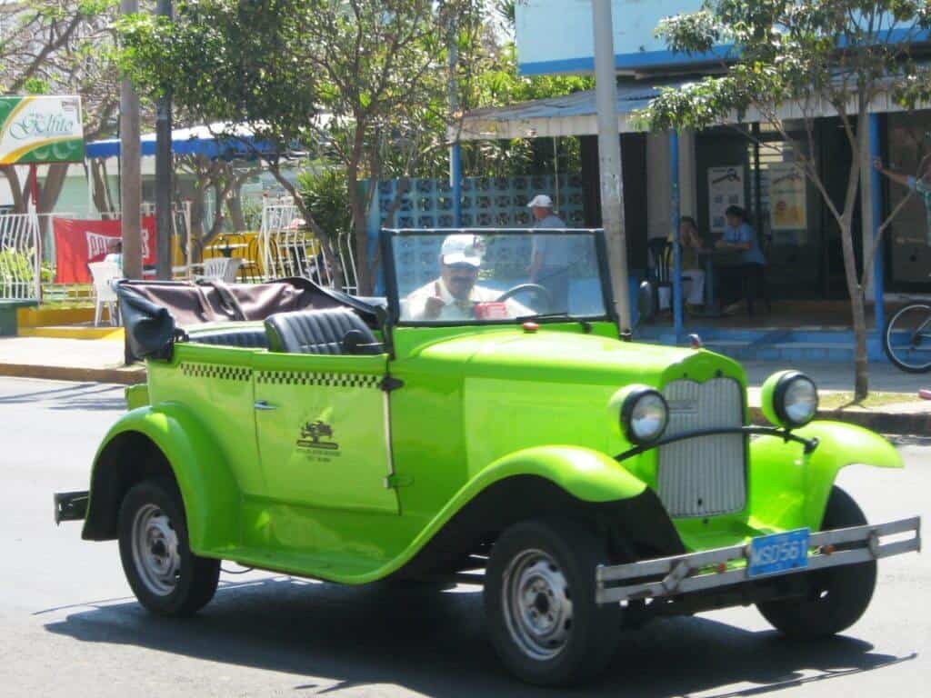 A bright green vintage convertible taxi with a senior driver, cruising along First Street in Varadero, Cuba. The scene captures a vibrant street atmosphere with outdoor cafés and pedestrians in the background, reflecting the lively and colorful urban life of Varadero.