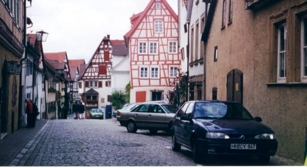 A quaint cobblestone street in Bad Wimpfen, Germany, lined with traditional half-timbered houses displaying rich red and white facades. Two cars are parked on the side, and a few pedestrians can be seen walking along the street. The architecture and the quiet atmosphere convey a sense of historical charm typical of German small towns.