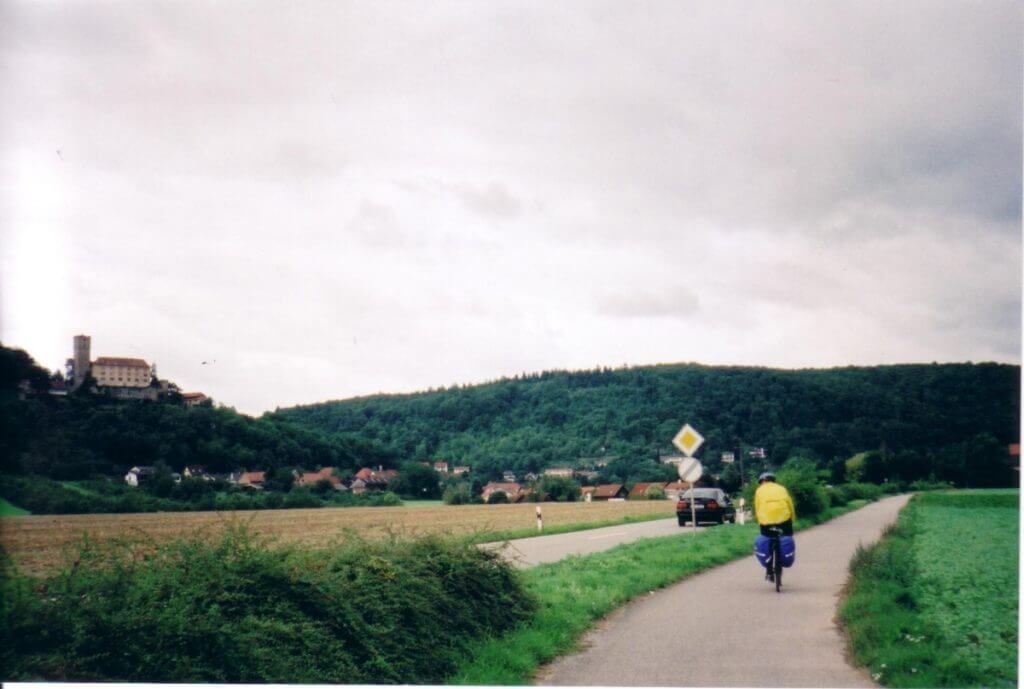 A cyclist in a yellow jacket is biking along a rural road on the Neckar Valley Cycling Route. In the background, a large, historical building sits atop a lush green hillside. The overcast sky adds a calm and serene feel to the scene.