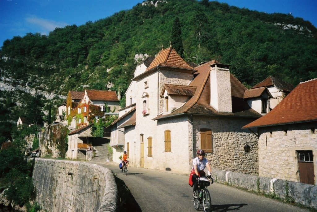 Cycling in Dordogne France on a ride through a picturesque French village along the Lot River, with charming traditional houses and a backdrop of lush green hills under a bright blue sky.