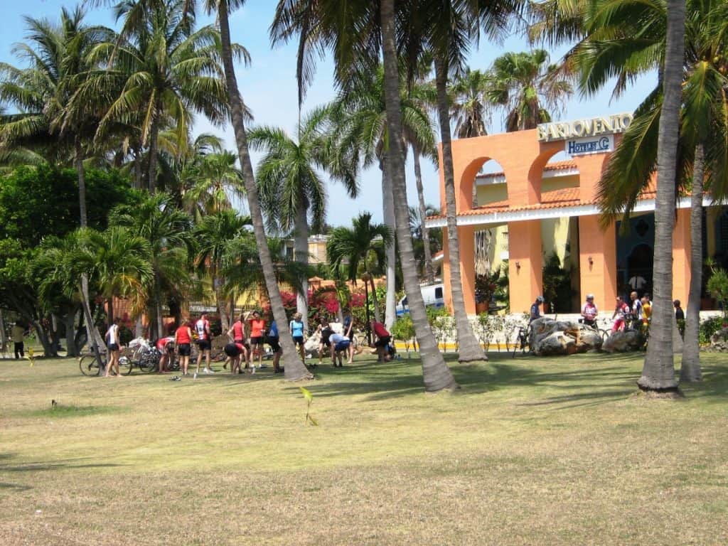 A group of cyclists, dressed in colorful attire, gather on a grassy area in front of the Varadero Hotel in Cuba, preparing for a ride. The hotel, featuring a distinctive orange façade and arched entryways, is framed by tall palm trees under a clear, sunny sky.