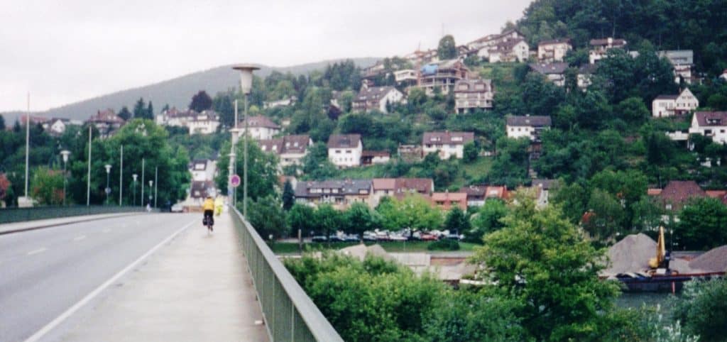 A cyclist approaches Eberbach on the Neckar Cycling Route, with the verdant hillside town in the background. The bridge offers a clear path into the town