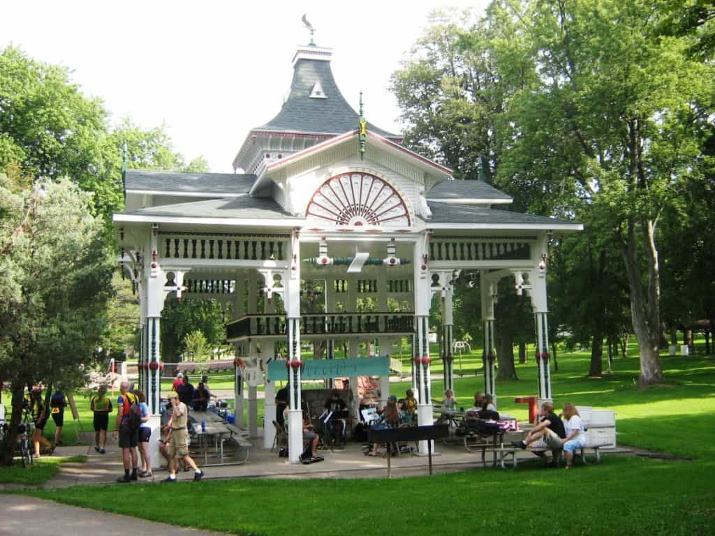 Cyclists relax and socialize around a classic Victorian-style gazebo in a park in Beaver Dam, Wisconsin. The gazebo, painted white with decorative green trim and a red fan-shaped detail at the peak, stands amidst lush green trees and well-kept lawns. People of various ages sit at picnic tables and on benches, enjoying a break in the shade on a sunny day.